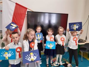 Uczniowie oddziału przedszkolnego w strojach galowych pozują do wspólnego zdjęcia. Dzieci maja przyczepiono kotyliony a w rękach trzymają wykonane przez siebie prace przedstawiające orły. W tle za nimi zza telewizora wystaje bokiem flaga Polski.