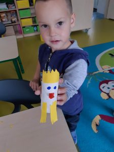 Uczeń oddziału przedszkolnego stoi przy stoliku. W ręku przed sobą trzyma wykonanego przez siebie z rolki orła z żółtą koroną i nóżkami.