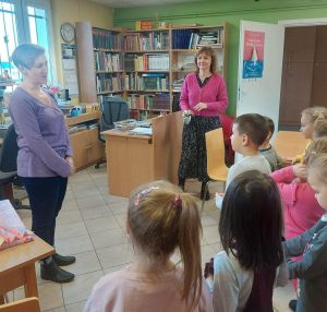 Pani Monika stoi na przeciwko uczniów oddziału przedszkolnego i do nich przemawia. Obok stoi Pani Alicja - wychowawczyni oddziału przedszkolnego. Zdjęcie wykonane w bibliotece w Ostaszewie.