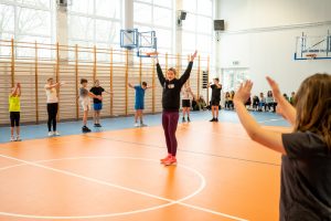 Pani Adrianna Sułek podnosi w górę ręcę. Uczniowie klas 5 i 6 ustawienie w dużym kole wokół nich również podnoszą do góry ręce.