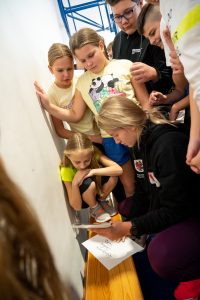 Adrianna Sułek rozdaje autografuy uczniom.. którzy ją otaczają. Lekkoatletka kuca przy ławce na której leży podpisana przez nią kartka.