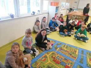 Uczniowie siedzą na podłodze i dywanie swojej klasy trzymając na kolanach pluszowe misie.