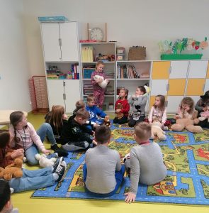 Uczniowie siedzą na brzegu dywanu trzymając swoje pluszowe misie. Jedna uczennica stoi trzymając w rękach białego misia.