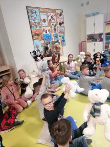 Uczniowie siedzą na podłodze swojej sali lekcyjnej. W górę podnoszą ręce z trzymanymi w nich pluszowymi misiami.