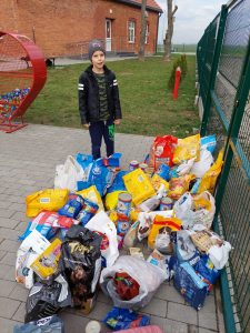 Przy bramie szkoły leżą ułożone dary dla zwierząt - karmy i puszki. Obokstoi jeden z uczniów.