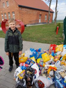 Zdjęcie ułożonych na szkolnym chodniku karm dla zwierząt. Obok nich stoi uśmiechający się chłopiec. W tle widać trawnik i fragment budynku szkoły.