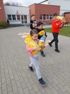 Zdjęcie uczniów idących szkolnym boiskiem i niesących w rękach karmy dla zwierząt. W tle widać budynek szkoły.