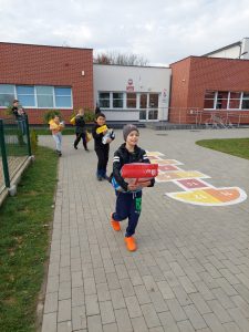 Zdjęcie uczniów idących szkolnym boiskiem i niesących w rękach karmy dla zwierząt. W tle widać budynek szkoły.