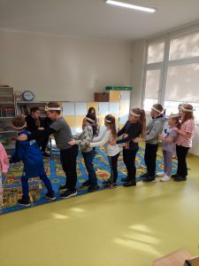 Dzieci stoją ustawione bokiem do zdjęcia w jednym rzędzie. Każdy uczeń ma ręce oparte na biodrach kolegi przed nim. Dzieci na głowach mają białe, papierowe opaski.