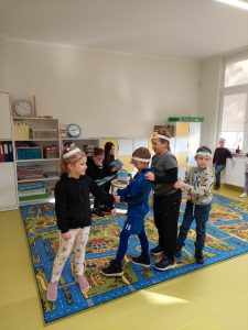 TRoje uczniów stoi bokiem do zdjęcia w jednym rzędzie opierając ręce na biodrach kolegi stojącego przed nim. Pierwszu uczeń trzyma za rękę dziewczynkę stojąca przodem do zdjęcia. Dzieci na glowach mają białe papierowe opaski. Zdjęcie wykonane w sali lekcyjnej z dywanem.