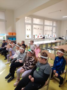 Dzieci siedzą na krzesełkach ustawionych obok siebie w dwóch rzędach. Dzieci na głowach mają białe papierowe opaski i patrzą przed siebie.