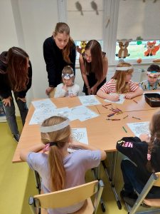 Uczniowie siedzą przy połączonych stolikach i kolorują kolorowanki warzyw. Nad nimi stoją trzy uczennice klasy ósmej i podziwiają ich pracę.