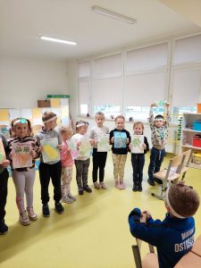 Dzieci stoją w jednym rzędzie trzymając w dłoniach kolorowe rysunki. Na przeciwko oparty o krzesło stoi jeden uczeń. Dzieci na głowach mają białe papierowe opaski.