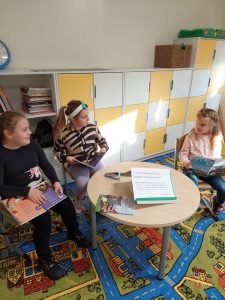 Obok okrągłego stolika na krzesełkach siedzą trzy uczennice. Dziewczynki spoglądają na siebie. Na kolanach mają rozłożone książki.