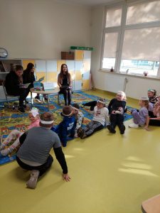 Na podłodze sali lekcyjnej siedzą uczniowie z białymi opaskami na głowach. Przed nimi na krzesełkach przy okrągłym stoliku siedzą trzy uczennice. dwie z nich trzymają na kolanach otwarte książki.