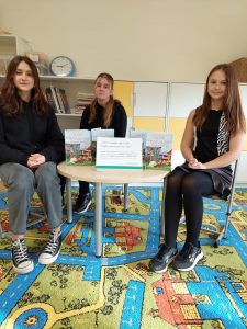 Trzy uczennice klasy ósmej siedzą na krzesełkach przy okrągłym stole z ustawionymi książkami i kartką z opisem akcji czytelniczej.