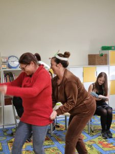 Dwie Panie stoją w rządku. Druga Pani trzyma pierwszą za sweter na wysokości bioder. Panie są radosne. W tle widać siedzącą na krzesełkku uczennicę z otwartą książką.