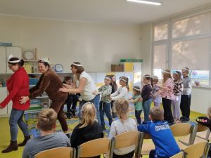 Trzy Panie oraz ustawione z nimi dzieci tworzą wężaopierając dlonie na biodrach osoby stojącej przed nimi. Wszyscy mają na głowach białe papierowe opaski. Przed nimi na krzesełkach siedzi czworo uczniów.