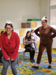 Dwie Panie z paiperowymi opaskami stoją na dywanie przyjmując pozy zwierzątek. Za nimi na krześle siedzi uczennica z otwartą książką.