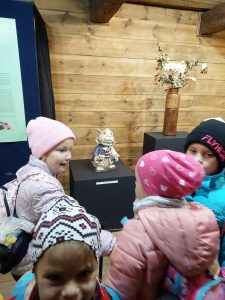 Dzieci stoją obok siebie naprzeciwko eksponatów muzeum etnograficznego. Przed uczniami stoi na podniesieniu figurka zwierzątka ubranego w strój oraz wazon z białymi kwiatami i jarzębiną.