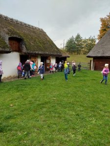 Uczniowie chodzą po polanie muzeum etnograficznego, na końcach której stoją zabudowania ze strzechą.