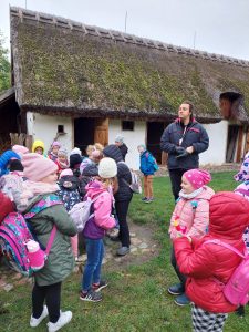 Uczniowie stoją na trawniku przed białym budynkiem ze strzechą na dachu. Przed nimi stoi Pan przewodnik z muzeum etnograficznego.