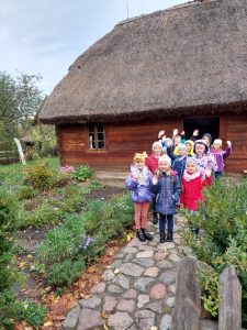 Uczniowie machają do zdjęcia. Ustawieni są w grupie na kamiennej ścieżce prowadzącej do drewnianego domu ze strzechą na dachu.