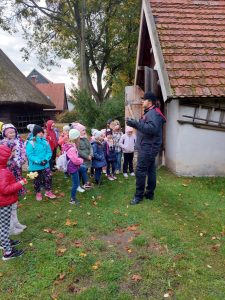 Uczniowie oddziału przedszkolnego stoją przed murowanym domkiem ustawionym na terenie Muzeum Etnograficznego. Obok domku stoi Pan przewodnik. Dzieci odwrócone są bokiem do zdjęcia.