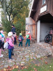 Uczniowie stoją przed budynkiem z otwartymi wrotami, za którymi widać fragment starodawnego powozu. Dzieci stojące najbliżej zaglądają do środka. Z boku stoi przewodnik muzeum etnograficznego.