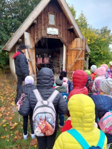 Uczniowie stoją przed drewnianym budynkiem z otwartymi wrotami, za którymi widać starodawny powóz. Obok budynku stoi ustawiony bokiem przewodnik muzeum etnograficznego.