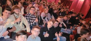 Uczniowie siedzą na czerwonych fotelach na auli. Część uczniów zauważyła zdjęcie i podnosi w góry kciuki.