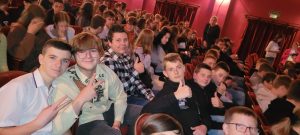 Uczniowie siedzą na czerwonych fotelach na auli. Część uczniów zauważyła zdjęcie i podnosi w góry kciuki.