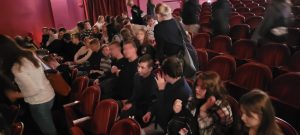 Uczniowie klasy ósmej podczas siadania na czerwonych fotelach w auli. Część uczniów już siedzi, część jeszcze wybiera miejsca.