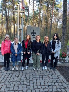 Członkowie szkolnego kola Wolontariatu - 6 dziewczynek, stoją w rzędzie obok siebie. W tle widać pomnik Harcerzy na Olku.