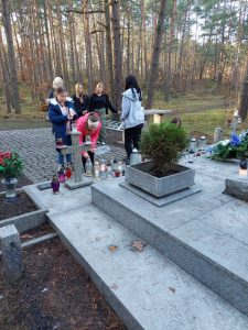 Członkowie szkolnego Koła Wolontariatu stoją przy pomniku Harcerzy na Olku. Jedna z dziewczyn pochyla się nas zniczem. Zdjęcie wykonane z oddali.