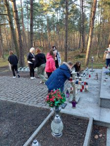 Członkowie szkolnego koła wolontariatu zapalają świeczki przy pomniku Harcerzy na Olku. Na zdjęciu widać fragment pomnika z ustawionymi na nim zniczami. Dwie uczennice pochylają się nad zniczami. W tle widać chodzących pozostałych uczniów.