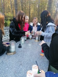 Członkinie szkolnego Koła Wolontariatu stoją przy płycie nagrobnej, na której ustawiono znicze. Dziewczynki po kolei je zapalają.