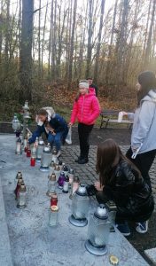 Członkinie szkolnego koła Wolontariatu pochylają się nad pomnikiem, przed którym ustawiają znicze. Obok stoją dwie uczennice.