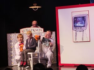 Zdjęcie sceny wykonane podczas przedstawienia. Na czarnym tle siedzi dziewczynka na białym krześle ze spuszczoną głową. Obok niej dwójka dorosłych ubranych na biało z pacynkami pluszowymi w dłoniach za nimi dekoracja przypomiajaca szuflady z napisem rejestracja, za którą stoi Pan. Z boku na białym tle z czerwoną ramką rysunek telewizora na którym wyświetla się usg.