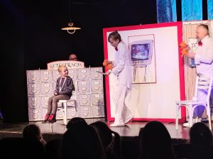 Zdjęcie sceny wykonane podczas przedstawienia. Na czarnym tle siedzi dziewczynka na białym krześle uśmiechnięta patrzy w bok. Po jej prawej stronie idzie dwójka dorosłych ubranych na biało z pacynkami pluszowymi w dłoniach. Za dziewczynką dekoracja przypominająca szuflady z napisem rejestracja, za którą stoi Pan. Z boku na białym tle z czerwoną ramką rysunek telewizora na którym wyświetla się usg.