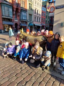 Uczniowie klasy pierwszej pozują do zdjęcia przy osiłku ustawionym przy toruńskim rynku. Część dzieci kuca przed osiolkiem a część za nim stoi.