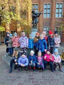 Uczniowie klasy pierwszej pozują przy fontanie z pomnikiem grającego na skrzypkach flisaka. Część dzieci kuca a część stoi.