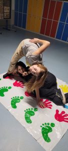 Dwie uczennice klasy ósmej podczas zabawy na macie z odciskami rąk i stóp. Jedna z dziewczyn leży na macie a druga nad nią się pochyla układając jedną rękę na macie.