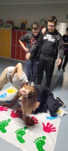 Dwie uczennice klasy ósmej podczas zabawy na macie z odciśniętymi kolorowymi dłoniami i stopami. Dziewczyny wyginają się w różnych pozach tak by utrzymać ręce i stopy na macie. Nad nimi stoi dwóch uczniów klasy ósmej i z zaciekawiem się przygląda.