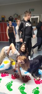 Dwie uczennice klasy ósmej podczas zabawy na macie z odciśniętymi kolorowymi dłoniami i stopami. Uczennice starają się utrzymać ręce i stopy na macie. W tle widać stojących innych uczniów klasy.