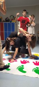 Dwie uczennice klasy ósmej podczas zabawy na macie z odciśniętymi kolorowymi dłoniami i stopami. Uczennice wyginają się tak by utrzymać ręce i stopy na macie. Za nimi stoi z podniesionymi rękami ich kolega z klasy.