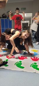 Dwie uczennice klasy ósmej podczas zabawy na macie z odciśniętymi kolorowymi dłoniami i stopami. Uczennice wyginają się tak by utrzymać ręce i stopy na macie. Za nimi stoi z podniesionymi rękami ich kolega z klasy.