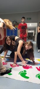 Dwie uczennice klasy ósmej podczas zabawy na macie z odciśniętymi kolorowymi dłoniami i stopami. Uczennice wyginają się tak by utrzymać ręce i stopy na macie. Za nimi stoi ich kolega przyglądający się całej sytuacji.