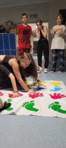 Dwie uczennice klasy ósmej podczas zabawy na macie z odciśniętymi kolorowymi dłoniami i stopami. Uczennice wyginają się tak by utrzymać ręce i stopy na macie. Za nimi stoi trójka innych uczniów a ich miny wskazują, że są wielce zadziwieni pozami koleżanek