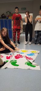 Dwie uczennice klasy ósmej podczas zabawy na macie z odciśniętymi kolorowymi dłoniami i stopami. Uczennice wyginają się tak by utrzymać ręce i stopy na macie. Za nimi stoi trójka innych uczniów a ich miny wskazują, że są wielce zadziwieni pozami koleżanek